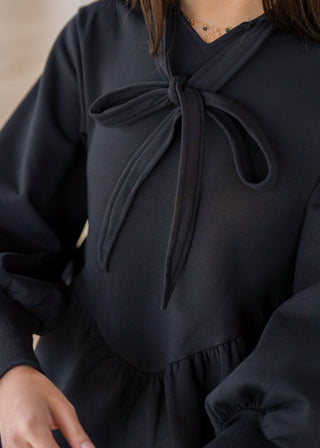 Person wearing a black dress with a bow detail sitting on a chair.